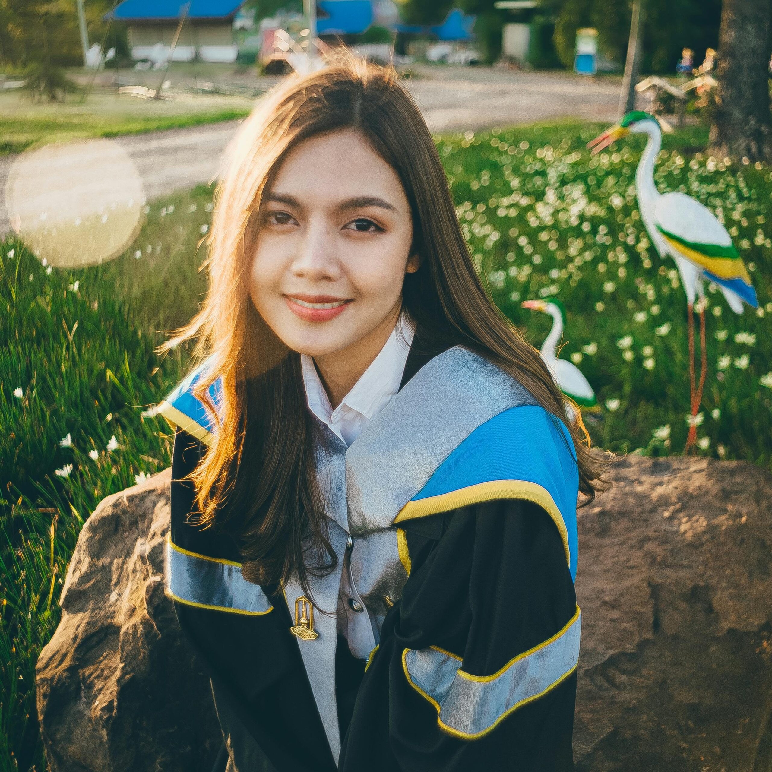 A young woman in graduation attire smiles outdoors in a serene park with decorative cranes.