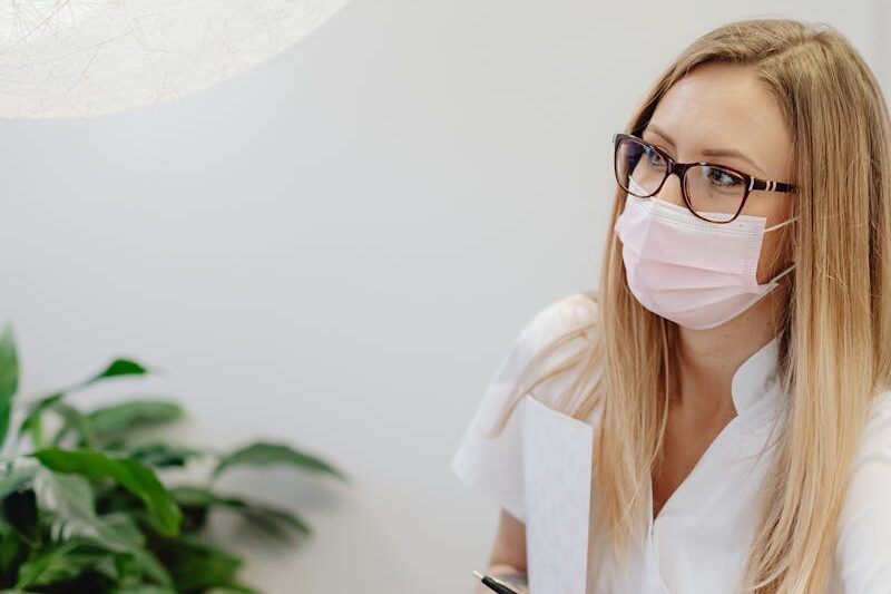 A female healthcare worker in a clinic wearing a face mask, standing at the reception desk.
